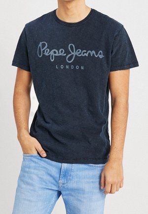 Mężczyzna ubrany w ciemnoniebieską koszulkę Pepe Jeans London i jasnoniebieskie dżinsy, z jedną ręką w kieszeni, na jednolitym tle.