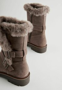 Bottes chelsea en suède marron avec bordure en fausse fourrure, double boucles et fermetures éclair sur le côté. Semelles texturées pour une meilleure adhérence, mettant en valeur des éléments de design robustes.