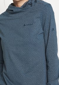 Dlouhý rukáv, modro-zelený top vyrobený z měkkého materiálu, s výstřihem ve tvaru cowl, jemn�ým puntíkovaným vzorem a malým logem na hrudi a rukávu.