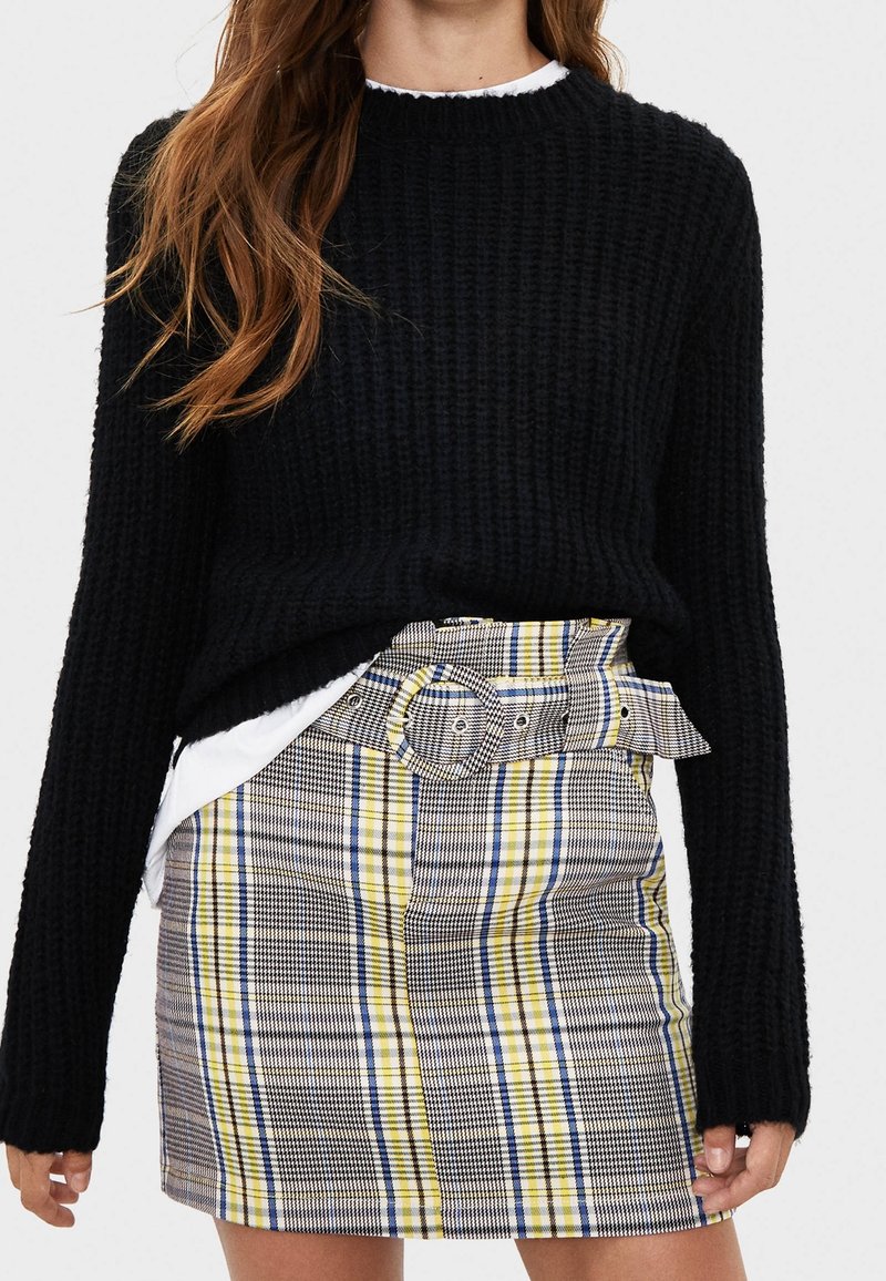 Černý pletený svetr s dlouhými rukávy, který je zkombinován s kostkovanou, vysokou mini sukni s žlutými, modrými a šedými vzory a páskem.