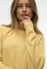 Gul sweater med tekstureret strik, ribbede manchetter og rund halsudskæring. Stoffet ser blødt og varmt ud og har et let splattet design.