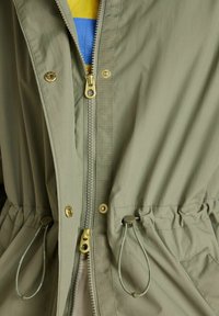 Geacă verde deschis cu două fermoare aurii, nasturi cu capse, șnururi ajustabile la talie și o vedere parțială a unei cămăși cu dungi galbene și albastre dedesubt.