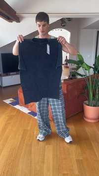 Una persona con pantalones de cuadros y zapatillas sostiene una gran camiseta oscura con una etiqueta de precio dentro de una sala de estar moderna.