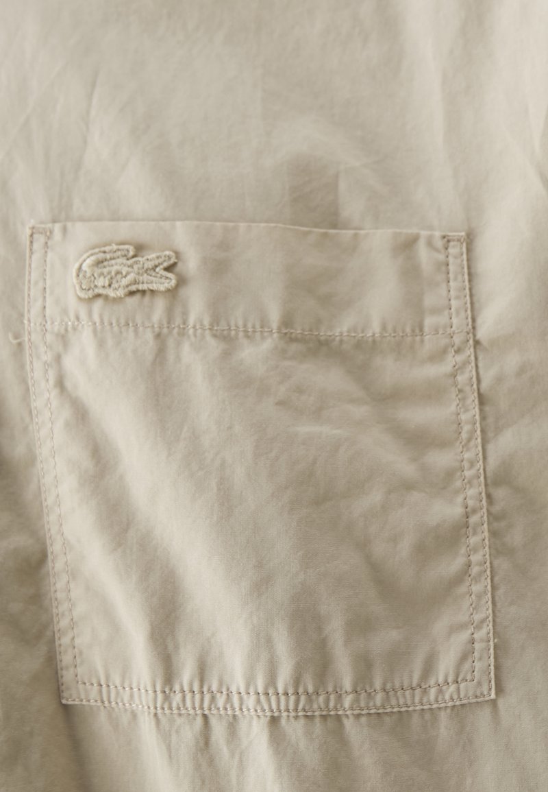 Beige stof met een gestructureerd oppervlak, met een klein wit geborduurd krokodillogo boven een paspelzak met gestikte randen.