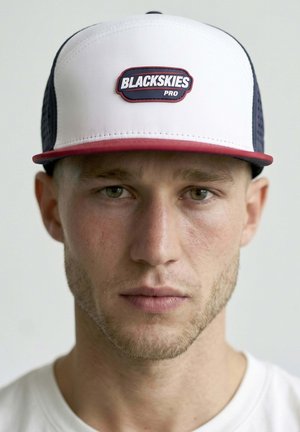 Man with light stubble wearing white and navy cap with red trim and "BLACKSKIES PRO" logo, neutral expression, and white shirt.