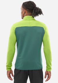 Mann mit geflochtenem Haar, der eine langärmelige, zweifarbige grüne Jacke und schwarze Hosen trägt, steht mit dem Rücken zur Kamera vor einem weißen Hintergrund.