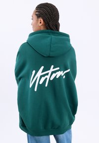 Zelena kapuca z velikim belim skriptnim logotipom na hrbtu. Ima prostoren kroj, kapuco s vezalkami ter rebraste manšete in spodnji rob.