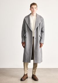 Γκρι παλτό από μαλλί με σχέδιο ψαροκόκκαλο, μεγάλους γιακάδες και διπλή σειρά κουμπιών, φορεμένο πάνω από εκρού ριμπ πλεκτό πουλόβερ και μπεζ παντελόνια. Καφέ μπότες.