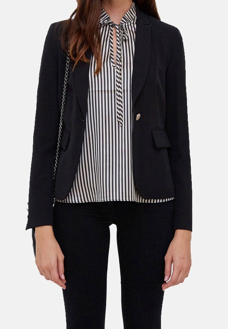 Femme portant un blazer noir sur un chemisier rayé vertical noir et blanc avec un col noué, assorti à un pantalon noir.