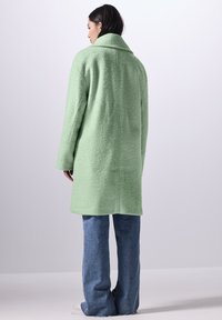 Cappotto verde texture con un ampio colletto, maniche lunghe e vestibilità rilassata, abbinato a jeans a zampa d'elefante blu chiaro e scarpe casual.