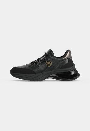 Sneakers sportive nere con suola spessa, linguetta metallica sul tallone, lacci e un ciondolo rotondo dorato con logo sul lato.