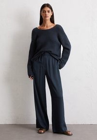 Donker marineblauw oversized gebreide trui met een brede halslijn, gecombineerd met losse, high-waisted broeken. Sandalen maken de outfit compleet.