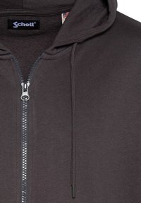 Haut de sweat à capuche zippé gris en tissu doux. Comprend une fermeture éclair sur le devant, des cordons de serrage, et une étiquette avec le nom de marque "Schott" visible sur le col.