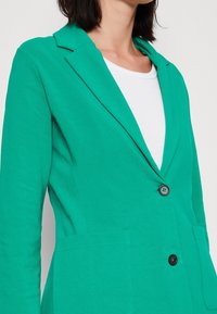 Blazer vert en tissu doux, présentant un design ajusté, des revers crantés, un bouton noir unique et deux poches avant.