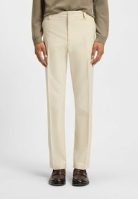Beige op maat gemaakte broek met een rechte pijp, vervaardigd uit een gladde stof. Voorzien van een sluiting met een knoop aan de voorkant en zijzakken. Gekozen in combinatie met donkere schoenen.