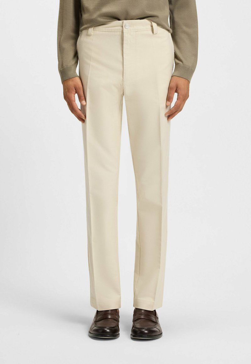 Beige op maat gemaakte broek met een rechte pijp, vervaardigd uit een gladde stof. Voorzien van een sluiting met een knoop aan de voorkant en zijzakken. Gekozen in combinatie met donkere schoenen.