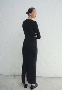 Dlouhé černé šaty z pletené tkaniny s kulatým výstřihem, dlouhými rukávy a rozparkem na zádech, doplněné černými botami a bílými ponožkami.
