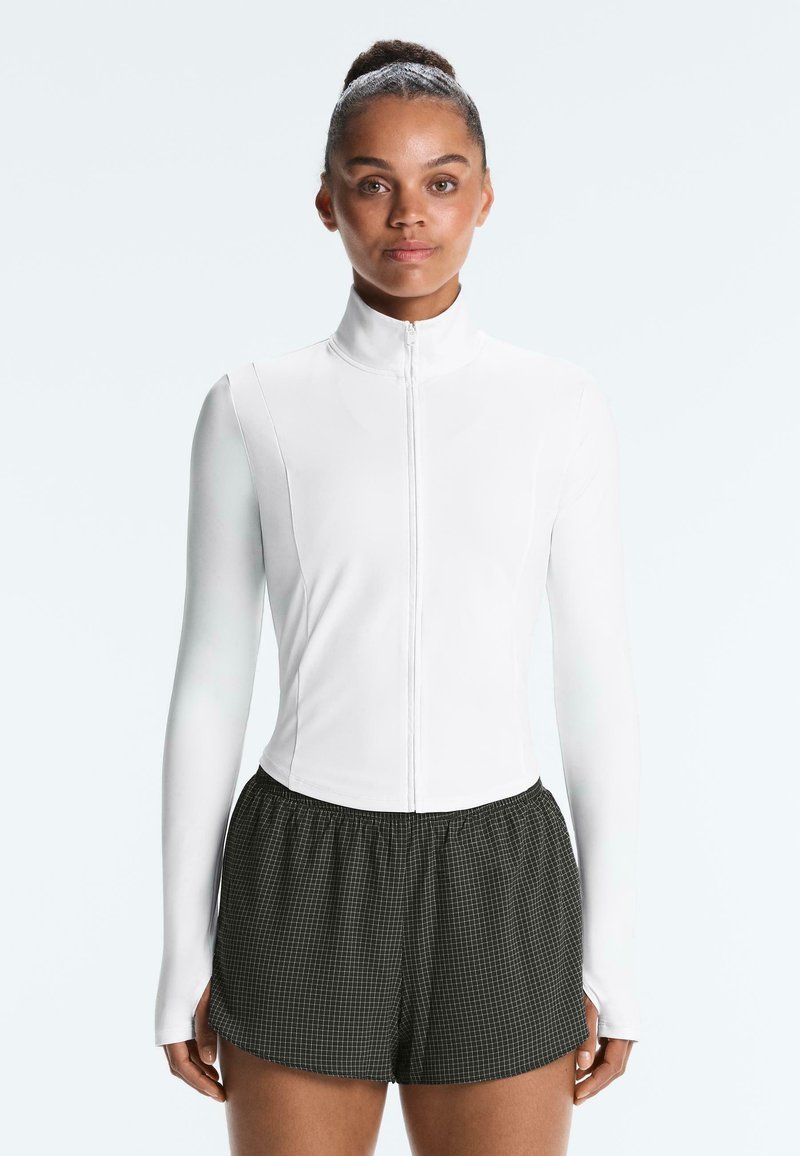 Femme portant une veste blanche zippée à manches longues et un short de sport noir à carreaux, debout face à un fond uni.