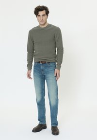 Chemise grise à manches longues, jean bleu délavé clair, ceinture en cuir marron et bottes marron. Le mannequin se tient les bras détendus le long du corps.