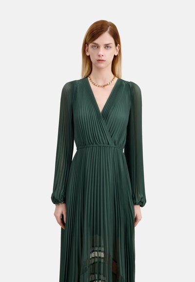 Długa, ciemnozielona, plisowana sukienka z dekoltem w kształcie litery V, długimi rękawami, talią podkreśloną paskiem i przezroczystym dolnym brzegiem. Uzupełniona złotym naszyjnikiem z łańcuszka.