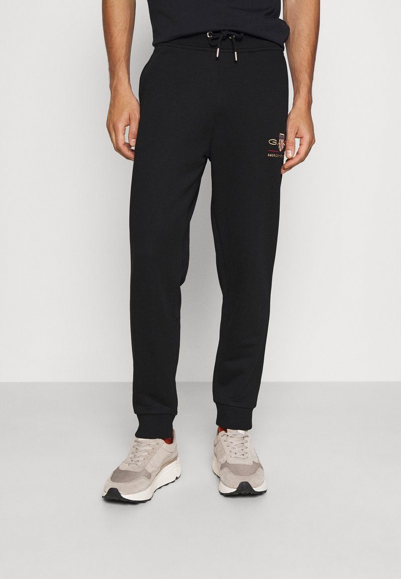 Zwarte sweatpants met een trekkoord in de taille en smal toelopende boorden. Bevat een klein gouden logo op de linker dij. Zacht, gemengd katoen materiaal.