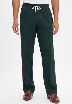 Mann trägt dunkelgrüne, locker sitzende Hose mit roten Seitenstreifen, weißem Kordelzugbund und braunen Tassel-Loafers auf weißem Hintergrund.