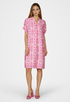Woman wearing knee-length pink and white geometric patterned dress with short sleeves and pink open-toe block heel shoes, standing.