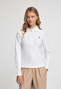 Vit långärmad polo t-shirt i bomull, med krage, knappstängning och en liten svart logotyp på vänster bröst.