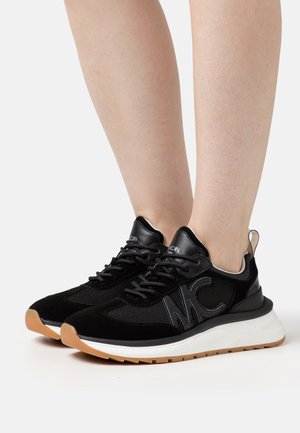 Beine, die schwarze und weiße klobige Turnschuhe mit braunen Sohlen tragen, die ein "MC"-Logo an der Seite und vorn Schnürsenkel haben.