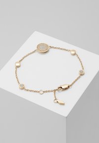 Pulsera de oro con una delicada cadena, un encantador redondo adornado con cristales y eslabones circulares lisos. Cierre de broche con una pequeña etiqueta.