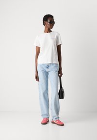 T-shirt bianco in cotone con logo, jeans leggeri a vita bassa blu chiaro, sneakers rosa con dettagli bianchi e una piccola borsa nera.