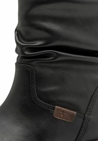 Cizmă înaltă din piele neagră, cu textură adunată, detalii subtile de cusătură și un mic etichetă logo maro ca accent. Finisaj exterior neted.