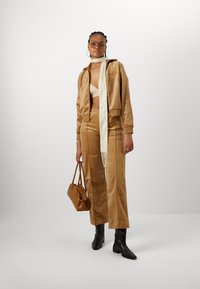Beige Satin-Trainingsanzug mit einer cropped, hochschließenden Jacke und weiteren Hosen, kombiniert mit einem cremfarbenen Schal und einer braunen Handtasche. Schwarze Stiefelettenschuhe runden den Look ab.