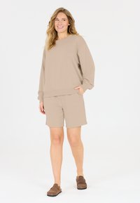 Beige tröja och shorts set i mjukt tyg, med en avslappnad passform, långa ärmar och sidofickor på shortsen. Bruna slip-on skor.