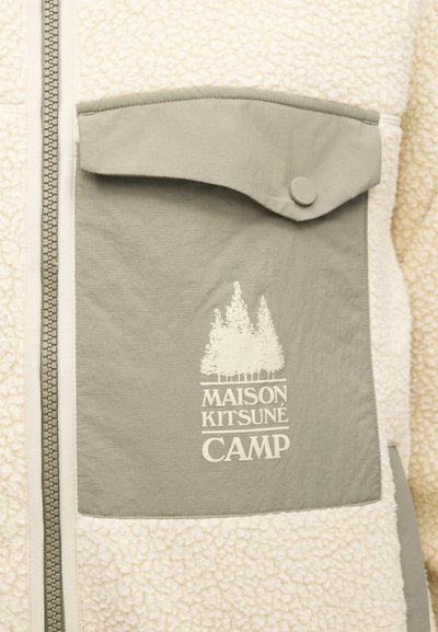 Stripete fleecejakke med en kremfarge tekstur, som har en grå lommeklaff med trykknapp og hvit trykket logo som leser "MAISON KITSUNÉ CAMP."