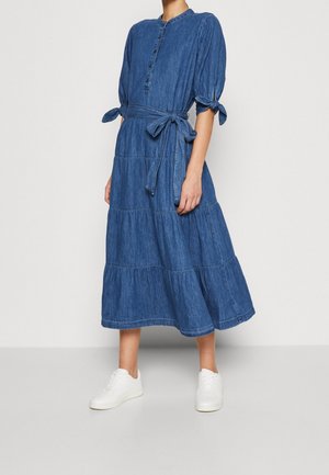 Nainen, joka käyttää sinistä denim-mittaista mekkoa, jossa on kyynärpäässä solmittavat pöyheät hihat, edessä nappilista ja vyö vyötäröllä, yhdistettynä valkoisiin lenkkareihin.
