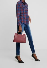 Bolso de cuero burdeos con asa superior redondeada, detalles en tono dorado y textura suave. Llevado con jeans azules y una camisa de cuadros.