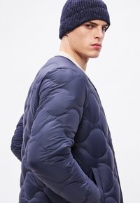 Ναυτικά μπλε μπουφάν με καπιτονέ σχέδιο, στρογγυλεμένες ενότητες, εμπρόσθιες τσέπες και κρεμ ριγωτό γιακά. Συνδυάζεται με σκούφο πλεκτό σε σκούρο χρώμα.