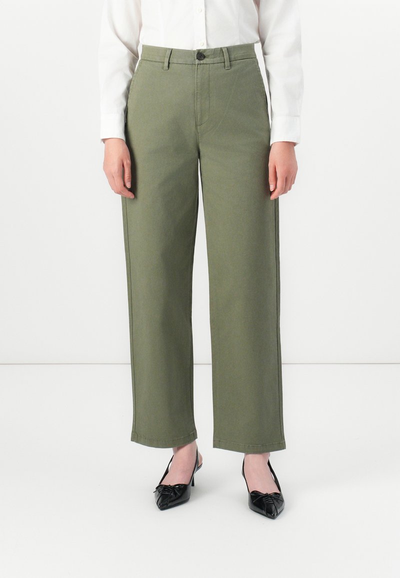 Donna che indossa pantaloni dritti verde oliva, camicia bianca a maniche lunghe con bottoni e tacchi a punta neri, in piedi su un pavimento bianco.