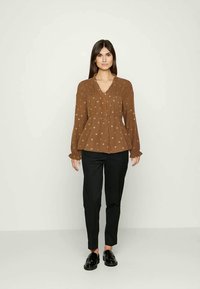 Blusa de mangas longas castanha, com padrão, e decote em V, com detalhes franzidos; combinada com calças pretas ajustadas e sapatos de loafers.