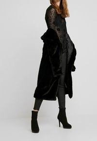 Abrigo de piel sintética negra con un diseño largo, combinado con un body floral negro transparente y pantalones negros ajustados. El calzado consiste en botas negras de tobillo.