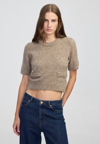 Młoda kobieta z długimi brązowymi włosami, ubrana w beżowy sweter z krótkim rękawem i ozdobami oraz ciemnoniebieskie dżinsy z wysokim stanem.