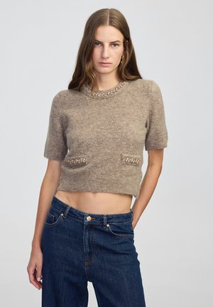 Młoda kobieta z długimi brązowymi włosami, ubrana w beżowy sweter z krótkim rękawem i ozdobami oraz ciemnoniebieskie dżinsy z wysokim stanem.