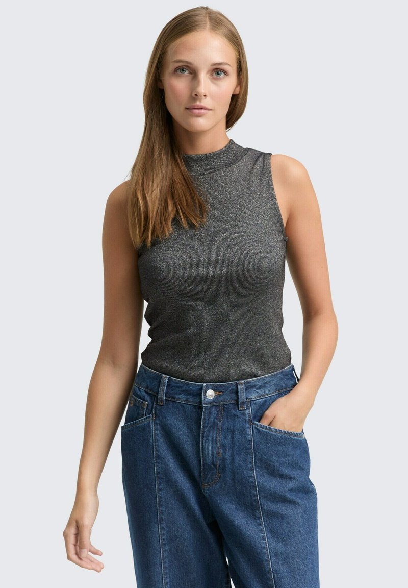 Ärmelloses, dunkelgraues, metallisches Oberteil mit hohem Ausschnitt, kombiniert mit locker sitzenden blauen Jeans. Das Model steht mit einer Hand in der Tasche.