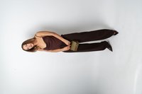 Hnedý tielkový top s prekladaným výstrihom, kombinovaný s tmavohnedými nohavicami s širokými nohavicami. Modelka drží olivovo zelenú kabelku s reliéfom. Špicaté opätky.