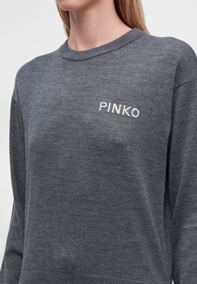 Жена, носеща тъмносив пуловер с дълги ръкави и "PINKO" бродирано в бяло в лявата част на гърдите.
