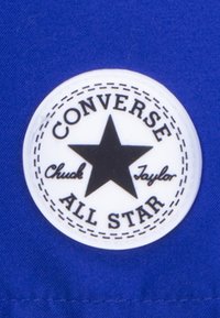 Okrogle belo logotip z črno zvezdo; besedilo pravi "CONVERSE ALL STAR" z "Chuck Taylor" spodaj, obrobljeno z prekinjeno črto. Ozadje je modro.