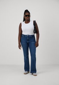 Top blanco sin mangas, jeans acampanados azules, zapatos grises, bolso negro. La modelo lleva gafas de sol negras. Tejido suave, diseño clásico de denim.
