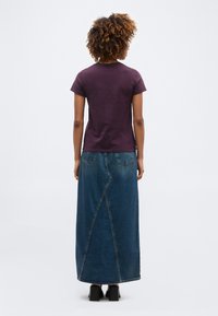 Γυναίκα με σγουρά μαλλιά, φοράει σκούρο μωβ μπλουζάκι με κοντά μανίκια, μακριά μπλε τζιν φούστα και μαύρα παπούτσια με τακούνι, στέκεται γυρισμένη με την πλάτη.