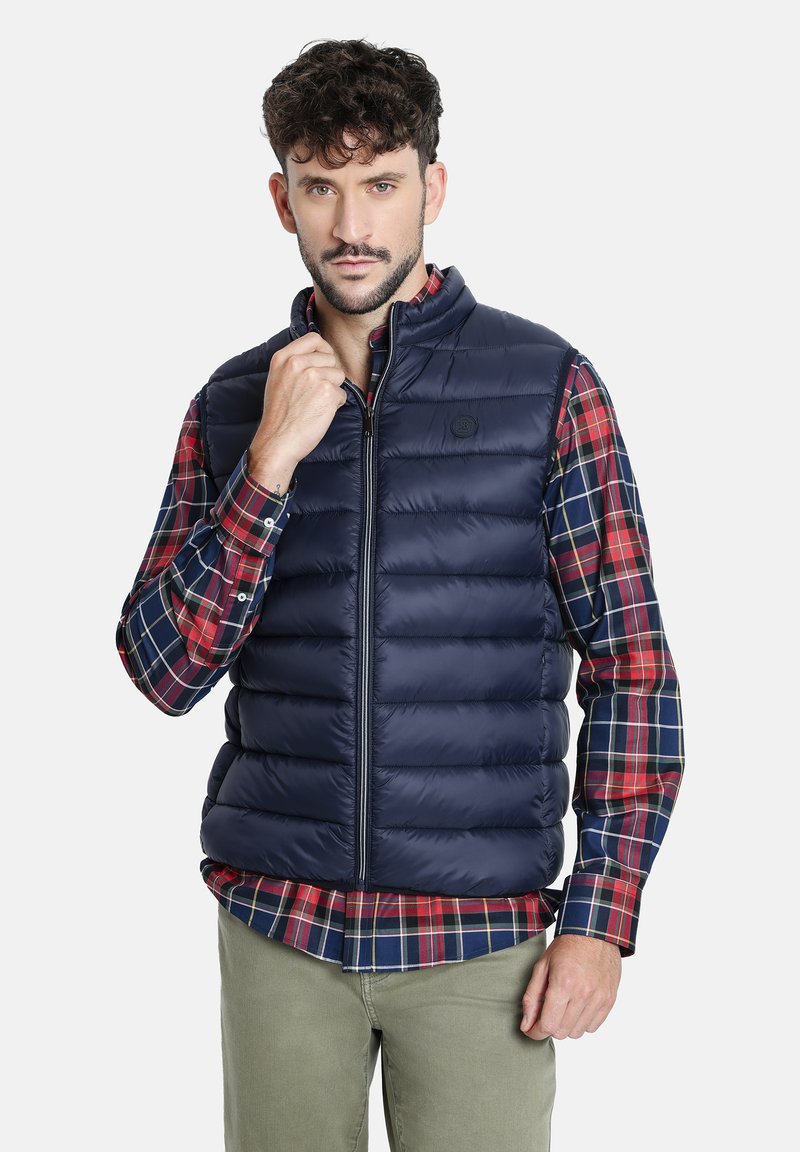 Chaleco acolchado navy con textura de rombos, cuello alto y cierre de cremallera. Lleva puesto sobre una camisa de cuadros en rojo y navy con base en navy liso.